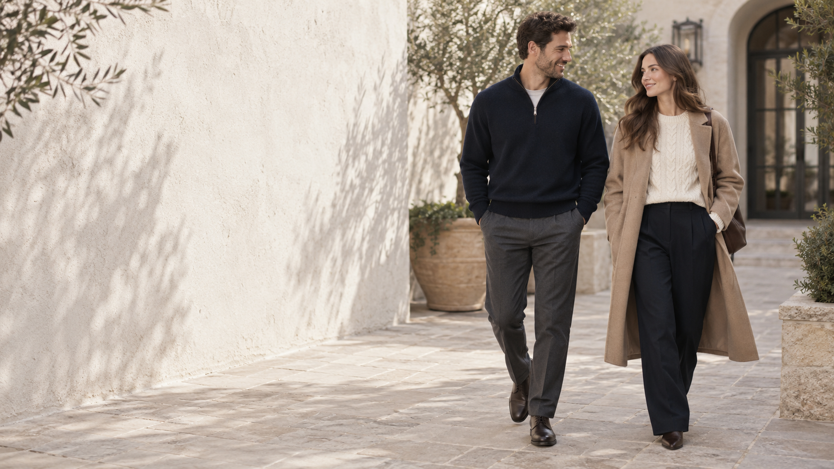 A man in a navy knit and a woman in an ivory sweater and camel coat walking in a courtyard
